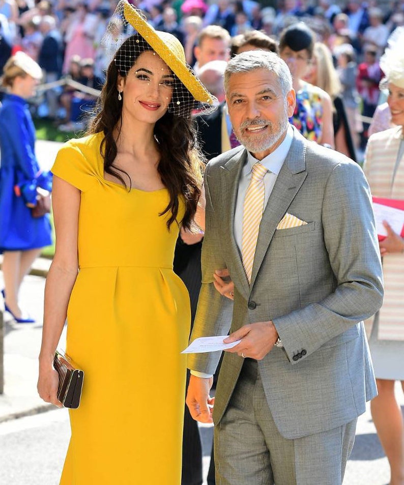May include: A couple poses for a photo. The woman is wearing a yellow dress and a wide-brimmed hat with a veil. The man is wearing a gray suit with a yellow and white striped tie.