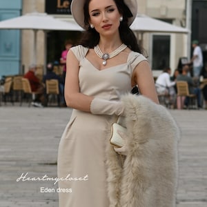 Pu&ograve; includere: Una donna indossa un abito beige con uno scialle di pelliccia bianca, un cappello bianco con un nastro nero e una collana di perle. Il testo "Heartmycloset Eden dress" &egrave; visibile nella parte inferiore dell'immagine.