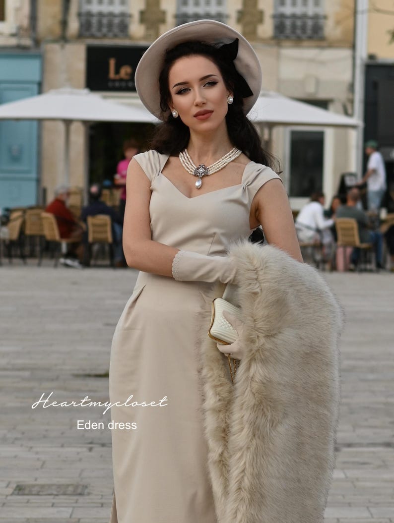 Pu&ograve; includere: Una donna indossa un abito beige con uno scialle di pelliccia bianca, un cappello bianco con un nastro nero e una collana di perle. Il testo "Heartmycloset Eden dress" &egrave; visibile nella parte inferiore dell'immagine.