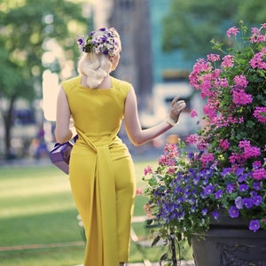 May include: A woman in a yellow dress with a flower crown in her hair walks away from the camera. She is carrying a purple purse and is wearing white gloves. The background is a blurry city street with a flower bed in the foreground.