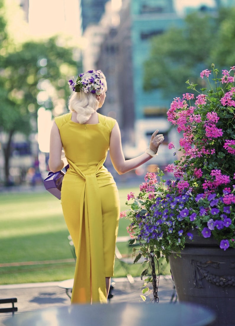 May include: A woman in a yellow dress with a flower crown in her hair walks away from the camera. She is carrying a purple purse and is wearing white gloves. The background is a blurry city street with a flower bed in the foreground.