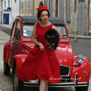 May include: A red vintage car with a woman in a red dress, hat, and heels. She is holding a small black dog. The car has a black roof and is parked on a cobblestone street. The text "heartmycloset" is visible.