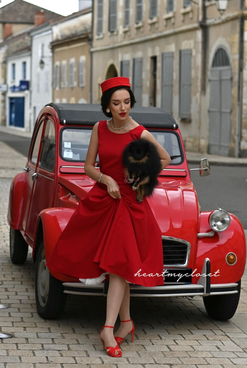 May include: A red vintage car with a woman in a red dress, hat, and heels. She is holding a small black dog. The car has a black roof and is parked on a cobblestone street. The text "heartmycloset" is visible.