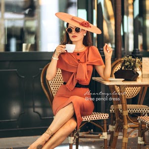 Puede incluir: Una mujer con un vestido lápiz color óxido y sombrero a juego, sentada en una mesa de café, sosteniendo una taza de café. El vestido presenta un detalle de lazo y un cinturón negro. La imagen incluye el texto "Heartmycloset Bree pencil dress".