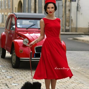 May include: A woman in a red dress and hat stands on a cobblestone street next to a vintage red car, holding a leash attached to a small black dog. The dress is knee-length and sleeveless.