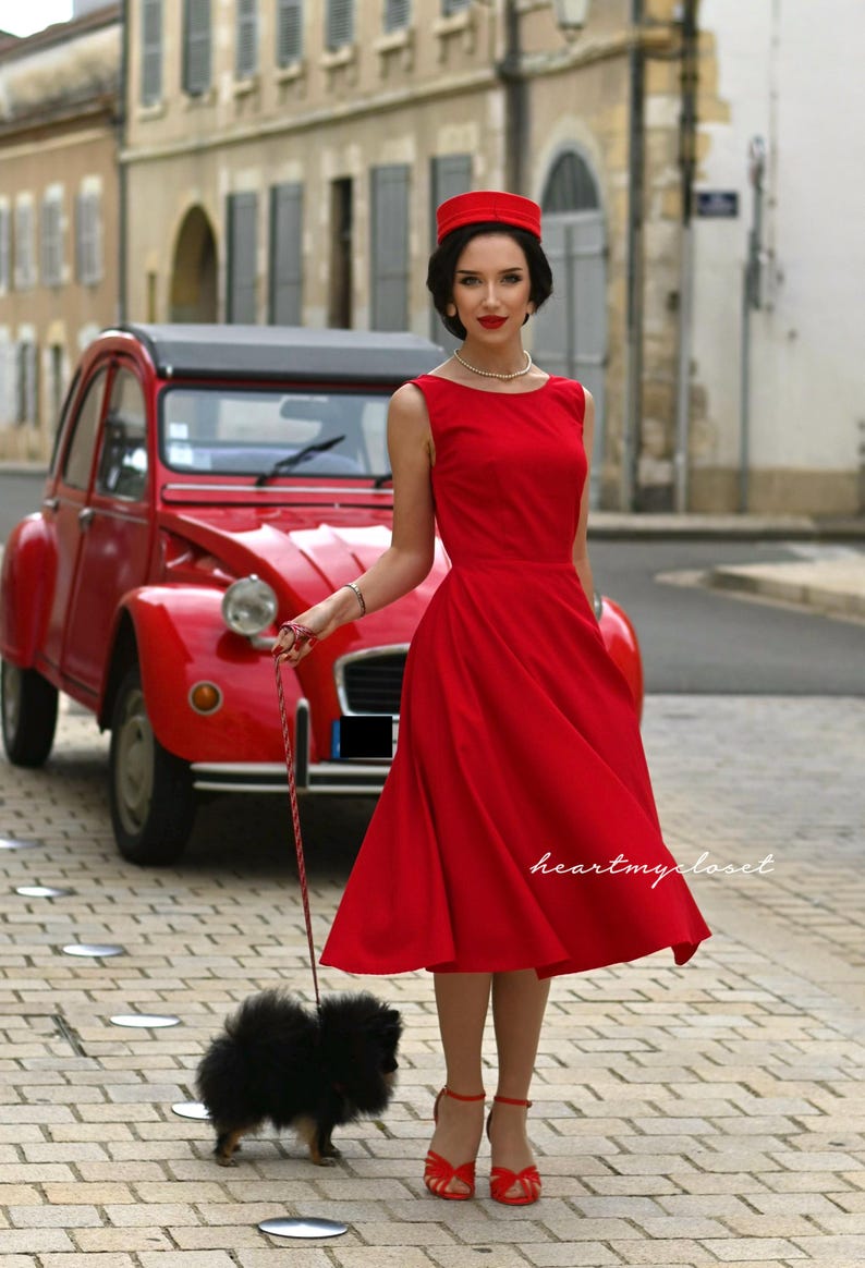 May include: A woman in a red dress and hat stands on a cobblestone street next to a vintage red car, holding a leash attached to a small black dog. The dress is knee-length and sleeveless.
