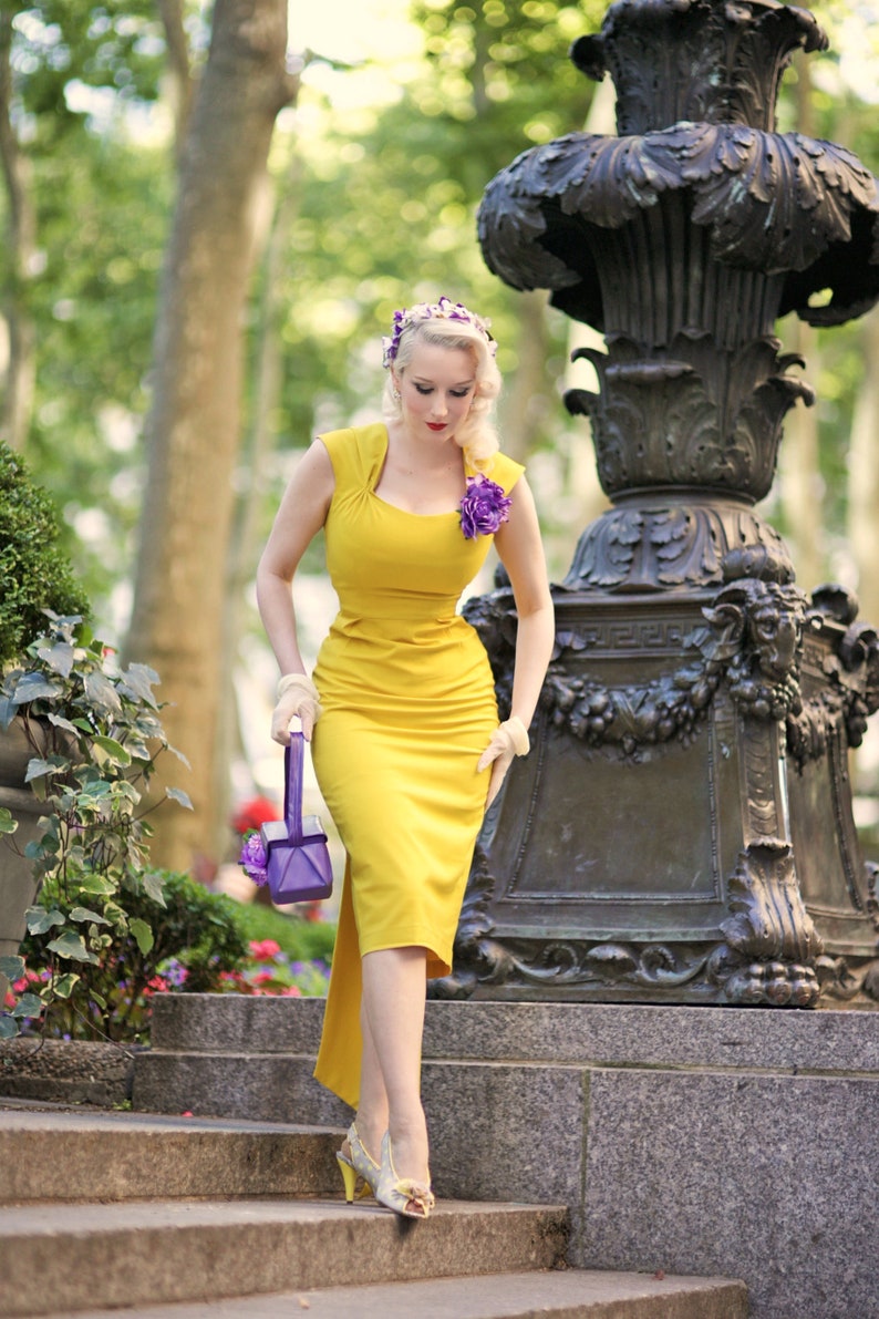 May include: A woman in a yellow dress with a purple flower on her shoulder walks down a set of stone steps. She is carrying a purple handbag with a flower detail.