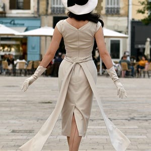 Pu&ograve; includere: Una donna indossa un abito beige con un grande fiocco bianco in vita. Indossa un cappello bianco e nero e guanti bianchi. L'abito ha una silhouette aderente e una gonna che le arriva alle ginocchia. Sta camminando su una strada acciottolata.