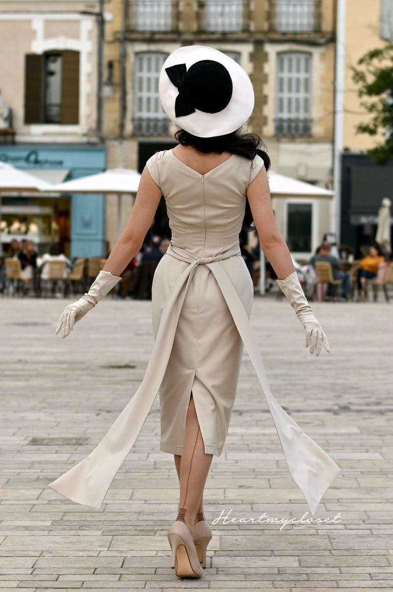 Pu&ograve; includere: Una donna indossa un abito beige con un grande fiocco bianco in vita. Indossa un cappello bianco e nero e guanti bianchi. L'abito ha una silhouette aderente e una gonna che le arriva alle ginocchia. Sta camminando su una strada acciottolata.