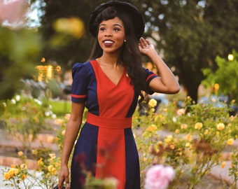 May include: A woman wearing a blue and red dress with a black hat and holding a vintage camera. She is standing in a garden with yellow flowers.