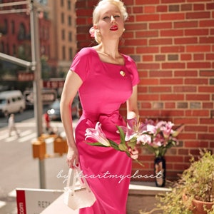May include: A woman in a pink dress with a white handbag and a bouquet of pink flowers. The dress has a square neckline and a bow at the waist. The woman is standing in front of a brick wall.