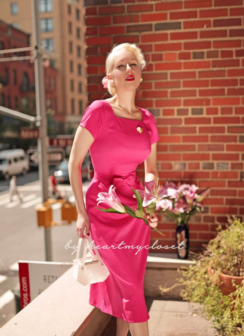 May include: A woman in a pink dress with a white handbag and a bouquet of pink flowers. The dress has a square neckline and a bow at the waist. The woman is standing in front of a brick wall.