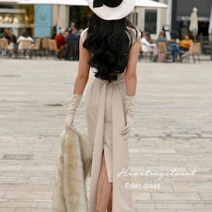 Pu&ograve; includere: Una donna indossa un abito beige con uno spacco alto, guanti bianchi e un cappello bianco e nero. Tiene una stola di pelliccia e cammina su una strada lastricata. Il testo "Heartmycloset Eden dress" &egrave; visibile nell'angolo in basso a destra dell'immagine.