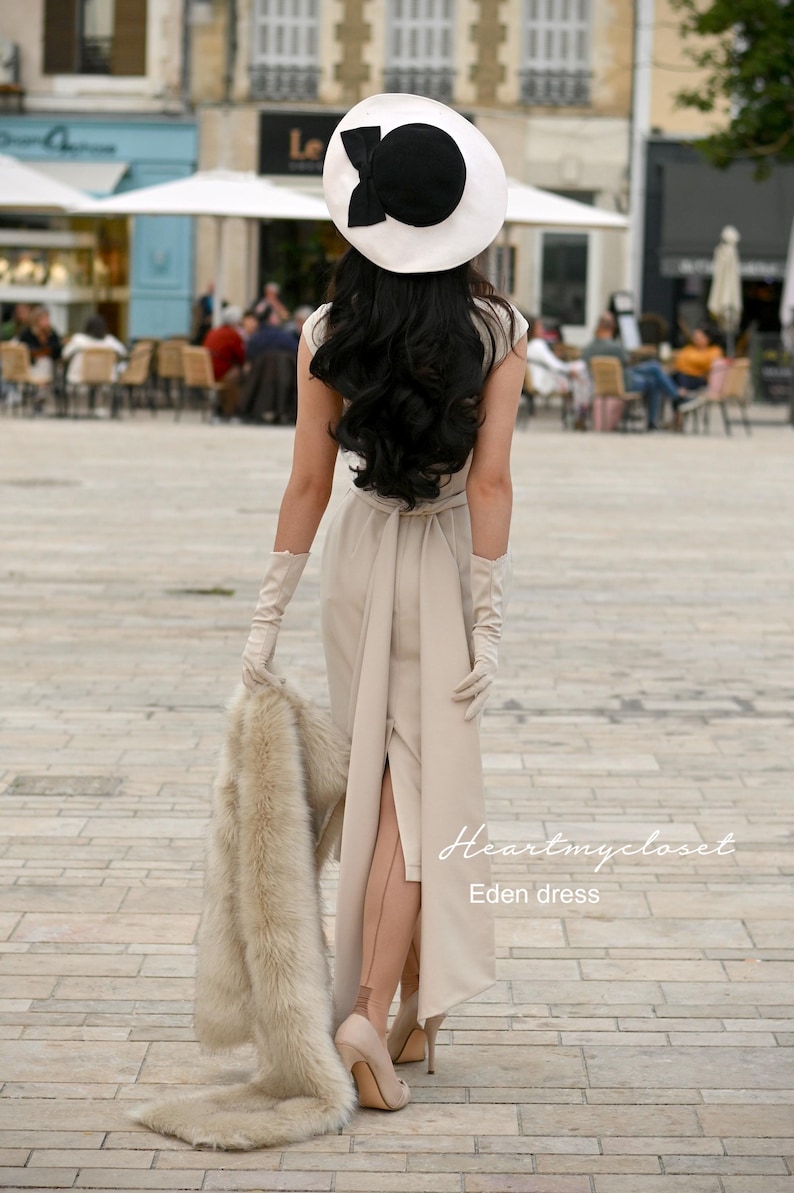 Pu&ograve; includere: Una donna indossa un abito beige con uno spacco alto, guanti bianchi e un cappello bianco e nero. Tiene una stola di pelliccia e cammina su una strada lastricata. Il testo "Heartmycloset Eden dress" &egrave; visibile nell'angolo in basso a destra dell'immagine.