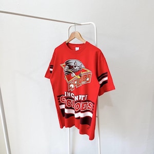 Può includere: T-shirt rossa con una grafica di un personaggio dei cartoni animati e la scritta "Cincinnati Cyclones". La maglietta ha strisce bianche e nere sulle maniche e sulla parte inferiore anteriore. La maglietta è appesa a un appendiabiti bianco.