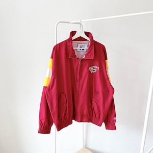 May include: A red zip-up jacket with yellow and white stripes on the sleeves. The jacket has a collar, two front pockets, and a logo patch on the chest. The jacket is hanging on a white hanger.