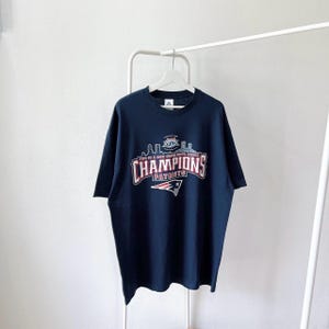 Könnte beinhalten: Marineblaues T-Shirt mit dem Text "CHAMPIONS PATRIOTS" in Rot und Weiß, mit einer Skyline-Grafik und einem Football-Logo. Das T-Shirt hängt an einem weißen Kleiderbügel.
