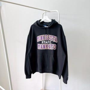 Peut inclure: Un sweat à capuche noir avec le texte MINNESOTA STATE MANKATO en lettres violettes à contours blancs sur la poitrine. Ce pull comprend une poche kangourou à l'avant, des manches longues à poignets côtelés et une capuche à cordon.