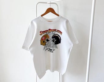 Puede incluir: Camiseta blanca con un gráfico de dos cascos de fútbol y el texto "Super Bowl XIX" y "Joe Robbie Stadium, Miami, FL Jan 19, 1985".