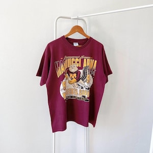 May include: A maroon t-shirt featuring a cartoon squirrel wearing a Minnesota Golden Gophers jersey and holding a hockey stick. The text "Inaugural Season Maucci Arena Minnesota Gophers That's the Ticket" is printed on the shirt.