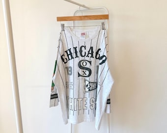 Puede incluir: Una camiseta blanca de manga larga con un patrón a rayas y un estampado gráfico en blanco y negro del logotipo de los Chicago White Sox. La camiseta tiene cuello redondo y está hecha de un material suave y cómodo.