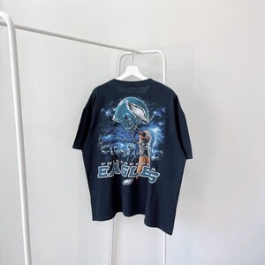Könnte beinhalten: Schwarzes T-Shirt mit einem Grafikdruck eines Footballspielers, der einen Helm der Philadelphia Eagles hält. Der Text "Philadelphia Eagles" ist unter dem Grafikdruck aufgedruckt.