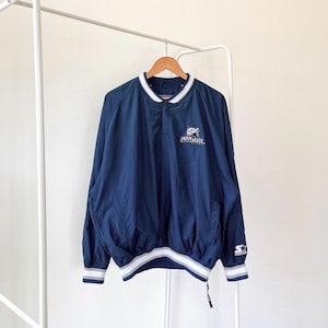 Puede incluir: Una chaqueta cortavientos azul marino con una banda blanca y gris a la cintura. La chaqueta tiene cierre de botones a presión y un logotipo blanco que dice "Penn State Nittany Lions". La chaqueta tiene un logotipo blanco "Starter" en la manga izquierda.