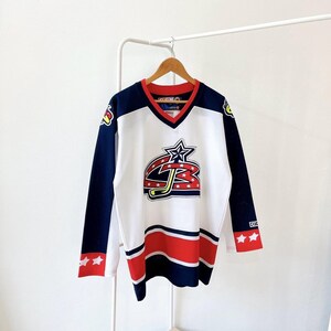 May include: White hockey jersey with navy blue sleeves and red accents. Features a star and hockey stick logo on the front. The jersey has a V-neck and is hanging on a wooden hanger.