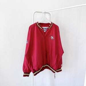 May include: Red and gold windbreaker jacket with a San Francisco 49ers logo on the front. The jacket has a snap button closure and a drawstring at the bottom.