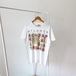 Può includere: T-shirt bianco con la scritta "ATLANTA CENTENNIAL OLYMPIC GAMES" in bordeaux. La maglietta presenta un collage di figure sportive olimpiche in varie pose. La maglietta è appesa a una gruccia di legno.
