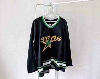 May include: Black hockey jersey with a green and white striped design at the bottom. The jersey has a V-neck collar with green trim and the word "STARS" in gold lettering with a green star logo.