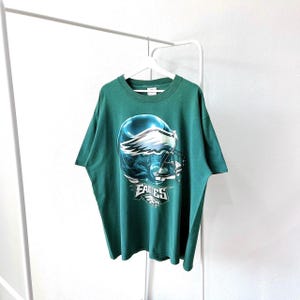 Könnte beinhalten: Grünes T-Shirt mit einem Aufdruck eines Footballhelms der Philadelphia Eagles und dem Wort "Eagles" auf der Vorderseite.