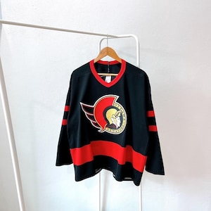 May include: Black hockey jersey with red and white accents. The jersey features the Ottawa Senators logo on the chest, a red V-neck, and red stripes on the sleeves and bottom. The jersey is hanging on a wooden hanger.
