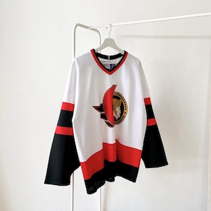 May include: White hockey jersey with black and red accents. The jersey features a V-neck collar with red trim and a team logo on the front. The sleeves are black with red stripes. The jersey is hanging on a white hanger.