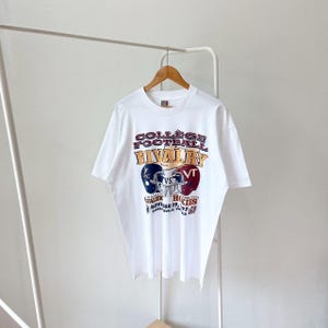 Könnte beinhalten: Weißes T-Shirt mit einem Vintage-Grafikdruck "College Football Rivalry". Das Design zeigt zwei Football-Helme, einen marineblauen und einen weinroten, mit dem Text "VT" und "Hokies". Das Shirt enthält auch das Datum "29. November 97".