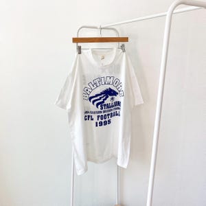May include: White t-shirt with a blue and white graphic print of a horse head with the text "Baltimore Stallions 1994 Eastern Division Champs CFL Football 1995".