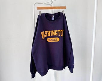 Op de afbeelding: Een donkerblauw sweatshirt met een gele afbeelding met de tekst "WASHINGTON HUSKIES".