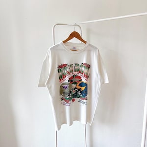 Pode incluir: Camiseta branca com um gráfico do Rose Bowl de 1992. O design inclui capacetes de futebol americano, o texto "Rose Bowl" e os nomes das equipes "Washington Huskies" e "Michigan Wolverines". A camiseta está pendurada em um cabide de madeira.