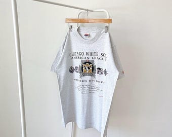 May include: Gray t-shirt with a vintage Chicago White Sox logo. The logo features a crest with a baseball, bats, and the text "Chicago White Sox, American League, Western Division".