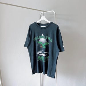 Op de afbeelding: Een donkergroen t-shirt met korte mouwen en de tekst Philadelphia Eagles in groene letters. De afbeelding bevat een groene adelaar die boven een wit driehoekig spotlight-effect vliegt en een donker schaduwsilhouet op de borst.