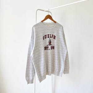 May include: A light grey sweatshirt with horizontal stripes. The word "GUELPH" is arched across the chest in maroon lettering, with a crest and the text "EST. 1864" below. The sweatshirt is hanging on a wooden hanger.