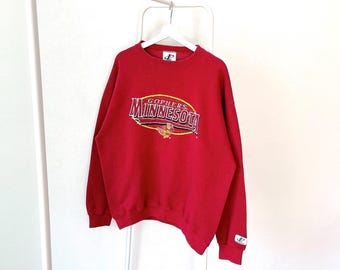 May include: Red sweatshirt with a white "Gophers Minnesota" logo in an oval shape with a gold outline. The logo also features a small gold mascot. The sweatshirt has a crew neck and long sleeves.