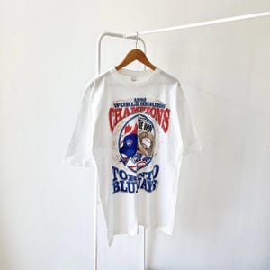 May include: White t-shirt with a graphic celebrating the 1993 World Series Champions, the Toronto Blue Jays. The design includes the team logo, a baseball glove, and the text "WE WIN". The shirt is displayed on a wooden hanger.