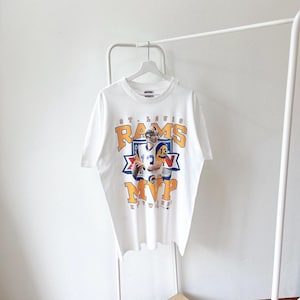 Puede incluir: Camiseta blanca con un gráfico de jugador de fútbol americano. El diseño incluye las palabras "St. Louis Rams" y "MVP" en naranja y azul. La camiseta está colgada en una percha blanca.