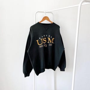 Könnte beinhalten: Schwarzer Sweatshirt mit einem goldenen gestickten Logo mit der Aufschrift "EAGLES USM EST. 1911".