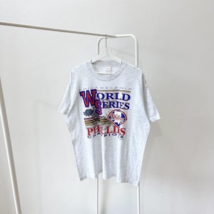 Puede incluir: Camiseta gris jaspeado con un diseño gráfico de los Philadelphia Phillies, campeones de la Serie Mundial. El diseño incluye el logotipo del equipo y las palabras "World Series" en rojo y azul.