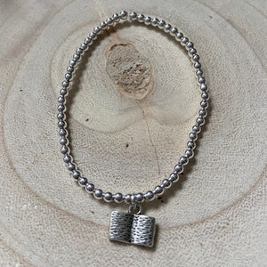May include: A silver beaded bracelet with a silver book charm. The bracelet is on a light brown wooden surface.