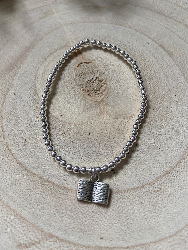 May include: A silver beaded bracelet with a silver book charm. The bracelet is on a light brown wooden surface.