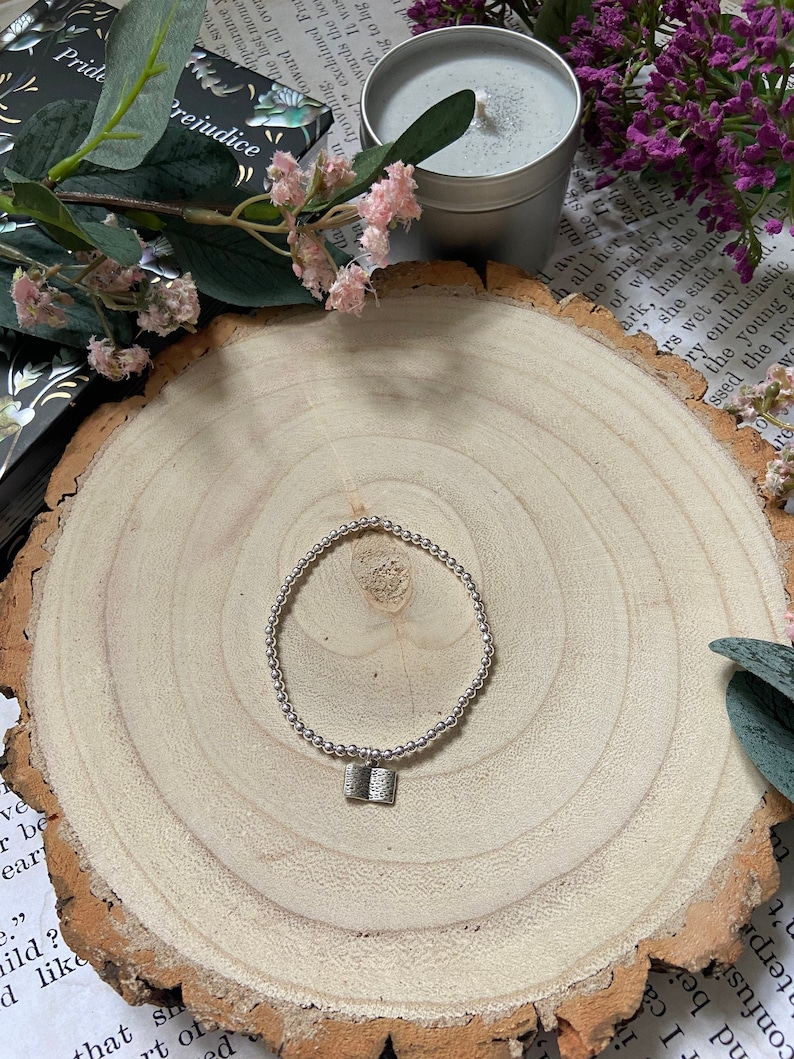 May include: A silver bracelet with a small book charm. The bracelet is on a wooden surface with a book and a candle in the background.