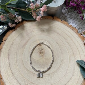 May include: A silver bracelet with a small book charm. The bracelet is on a wooden surface with a book and flowers in the background.
